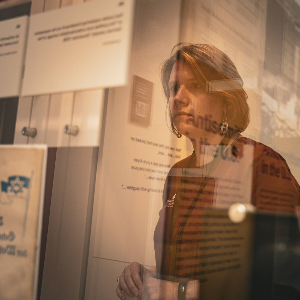 A woman behind a glass with "Antisemitism in the US" reflected upon it reading a museum description plaque.